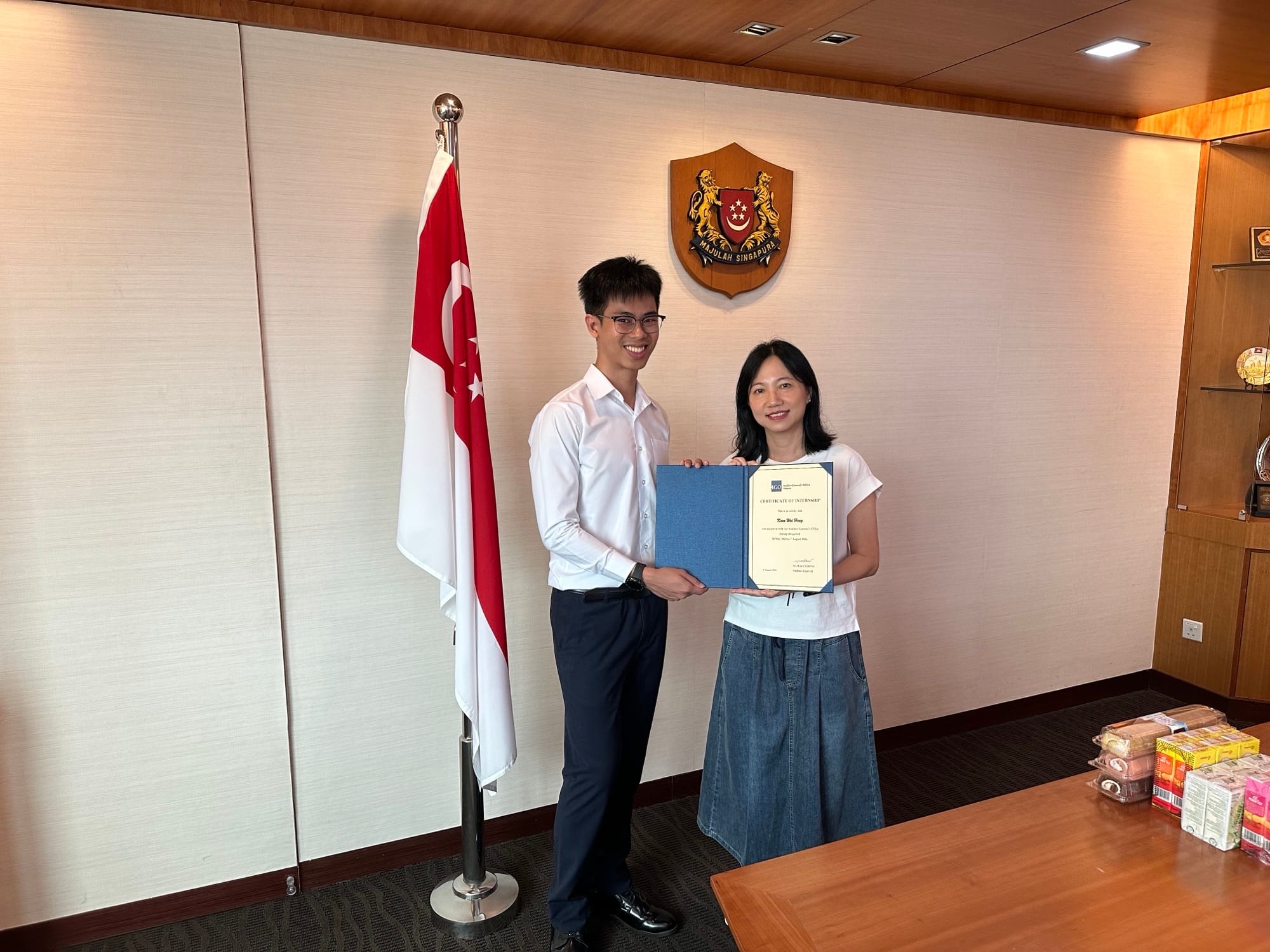 Two individuals are standing in front of a wall with the Singapore crest and flag, and two people holding a certificate.